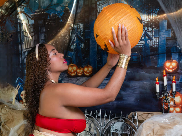 AishaRhodes with curly brown hair, wearing a red strapless top and gold bracelet, holds an orange pumpkin with a black jack-o'-lantern face, looking up at it.