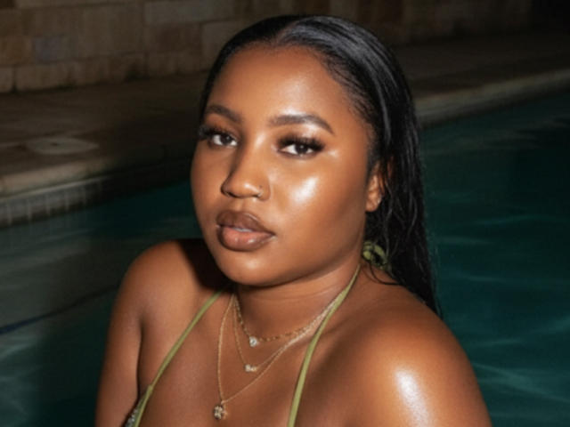 StarHottie, a dark-skinned woman with wet black hair, stands in a pool. She wears a green halter top, multiple thin gold necklaces. Her makeup is bold with shimmery eyeshadow and glossy lips. Her expression is neutral. The water is dark green, pool edge is tiled. StarHottie's skin is shiny, highlighting her facial features. The image is a photograph with a realistic style, focusing on her upper body.
