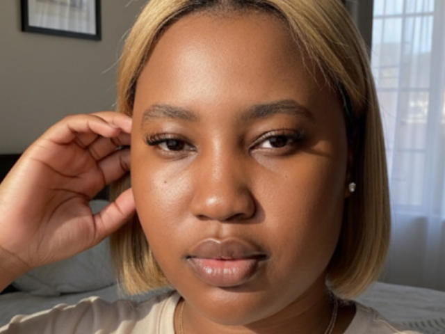 Close-up of StarHottie with medium brown skin, straight blonde bob hairstyle, and small diamond earring. StarHottie has arched eyebrows, brown eyes with subtle eyeliner, and full, slightly glossy lips. She's touching her right ear with her right hand, wearing a beige top. The image is well-lit with natural light from a window on the right. The texture of her skin is smooth with a small beauty mark on her left cheek.