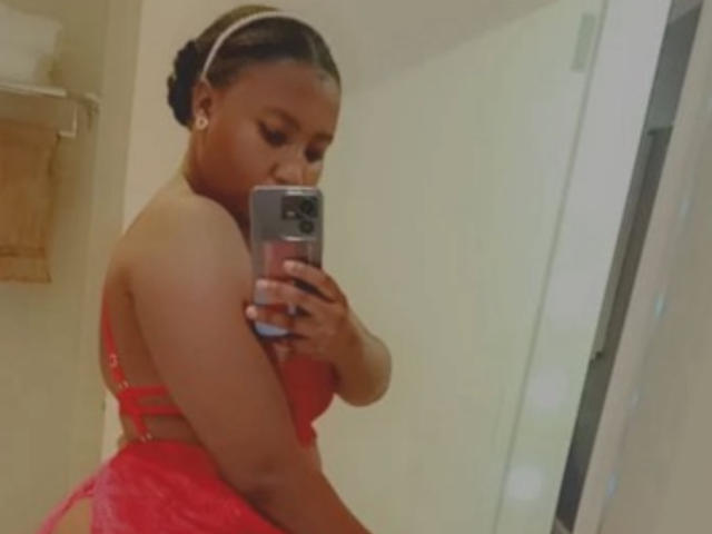 Eine attraktive Frau mit dunkler Haut, die rote Spitzendessousiers trägt, ihr Gesäß zeigt und ein Selfie vor einem Spiegel mit ihrem Handy macht, ihre Haare sind zu einem Dutt gebunden und sie trägt ein weißes Stirnband..