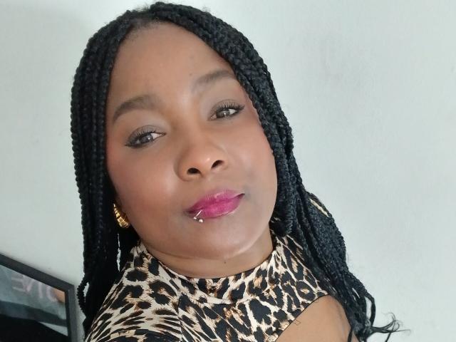 DakotaScoth with dark braided hair, brown skin, pink lipstick, gold earrings, and a leopard print top, looking at the camera with a slightly pouty expression.