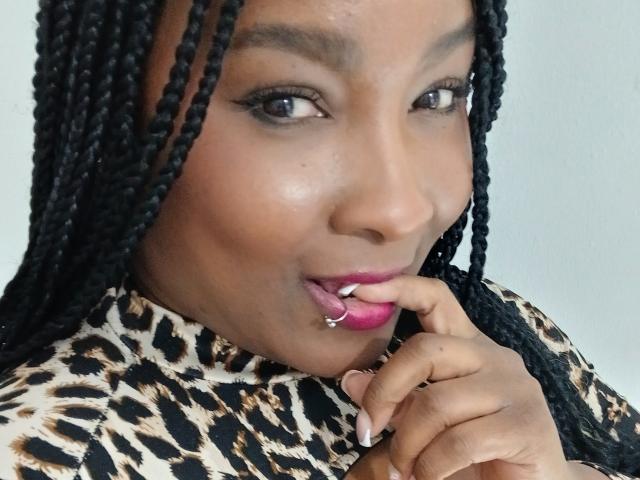 DakotaScoth with dark braids, brown skin, and pink lipstick, sucking on a silver ringed finger, wearing a leopard print top, looking at the camera.