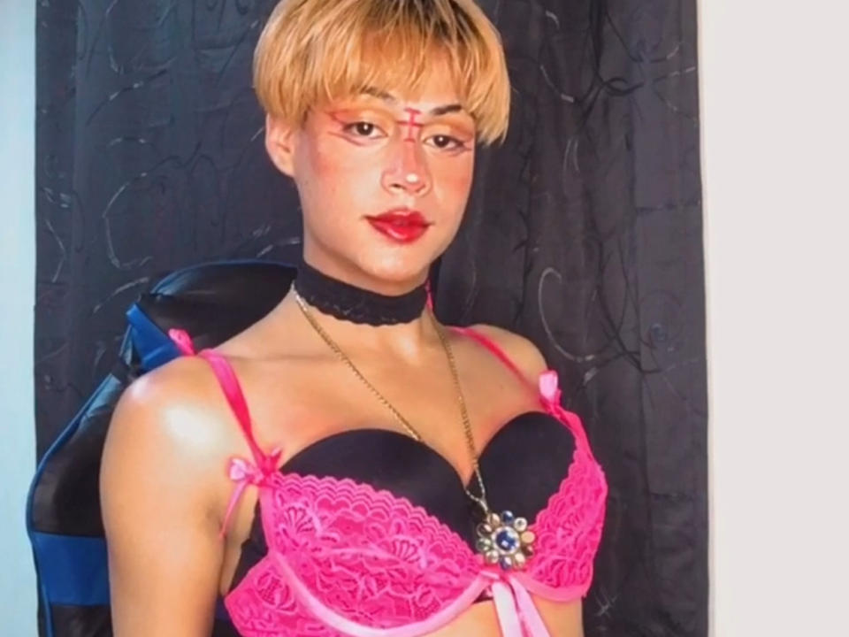 MachiSex has short, blonde hair and wears red face paint in a geometric pattern. They have light brown skin and red lipstick. MachiSex wears a black choker, a pink lace bra with black cups, and a gold necklace with a blue flower pendant. The bra has pink ribbons on the straps. The image is brightly lit, highlighting their smooth skin and confident expression. The bra's lace texture contrasts with the smooth skin of MachiSex's chest. The overall style is bold and provocative.