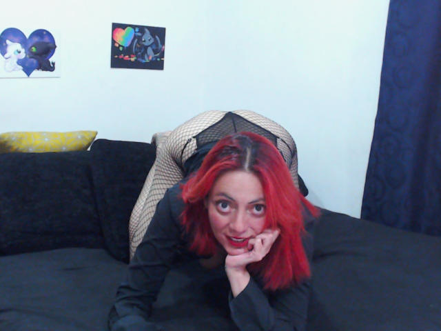 SamanthaSwan with bright red hair leans forward on a black couch, her face resting on her right hand. She wears a black long-sleeve shirt and fishnet pants, looking directly at the camera with a slight smile. Her red hair contrasts with her pale skin and dark clothing. Two small, colorful posters are on the white wall behind her. The couch has a yellow pillow on the left. The image has a casual, home setting.