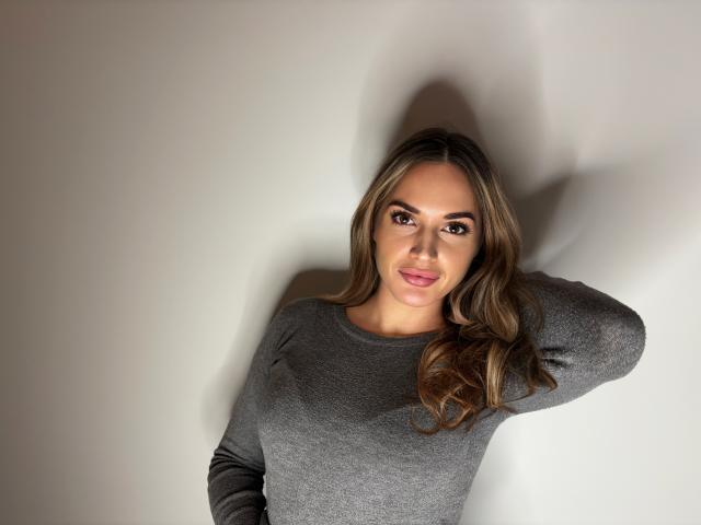 EmmaSlim with long brown hair, wearing a gray long-sleeve shirt, has her hand behind her head, smiling with pink lips, brown eyes, and full eyebrows.