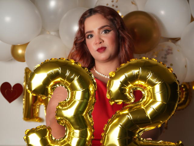DannaXFerluccio mit lockigem, rotem Haar, die glänzende, goldene "2"-Ballons hält, ein rotes Kleid und eine Perlenkette trägt und mit rotem Lippenstift in die Kamera schaut..