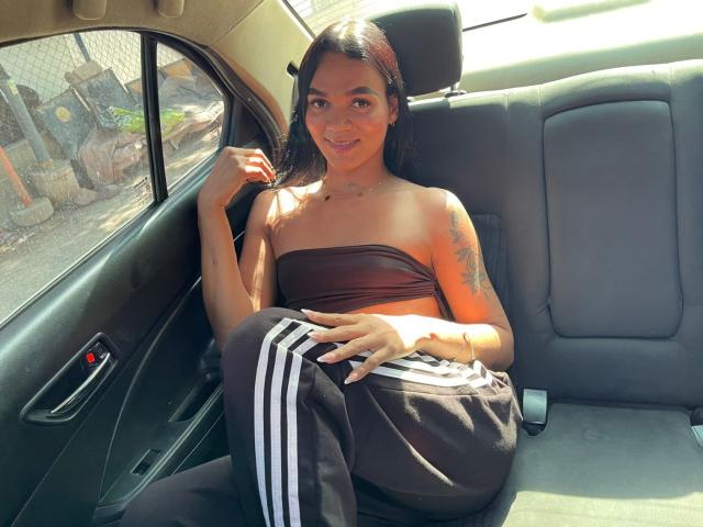 DulceDanesa, tan-skinned, with long dark hair, sits in a car, wearing a brown strapless top and black track pants with white stripes, smiling, tattoo on right arm.