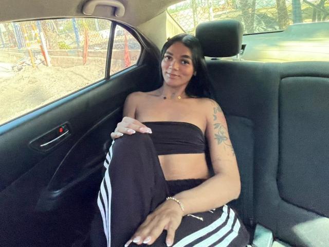 DulceDanesa with medium tits, black strapless top, black pants with white stripes, sitting in car, arm tattoo, smiling, dark hair, tan skin, bracelet, sunlight through window.