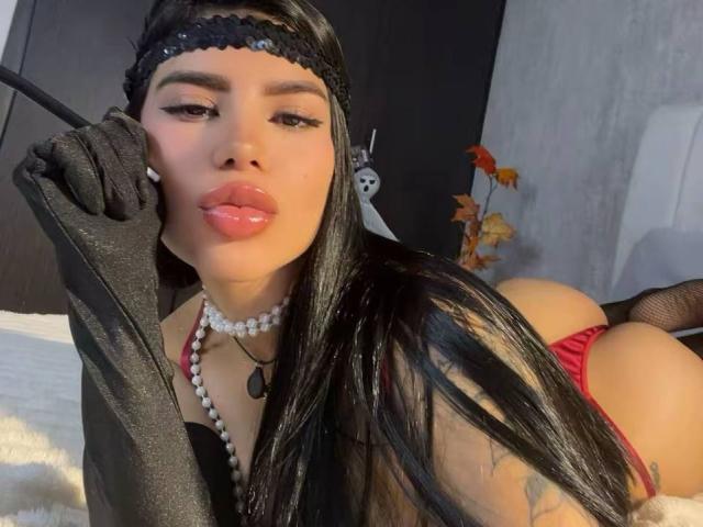 NikolRyder, con cabello negro largo, usando una diadema negra, guantes negros, un collar de perlas, una tanga roja y una blusa negra, está acostada en una cama, sosteniendo un látigo negro, con labios brillantes..
