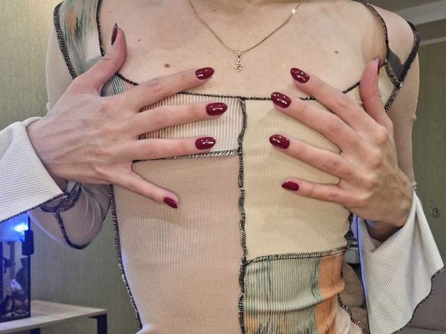 MilaYanis with hands covering her tits, wearing a sheer, ribbed, patterned dress, red glossy nails, thin gold necklace, in a dimly lit room.