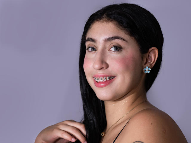 ErikaDavies has black hair, brown eyes, and a pinkish blush on her cheeks, wearing blue flower earrings and a gold necklace, smiling with her teeth slightly visible.