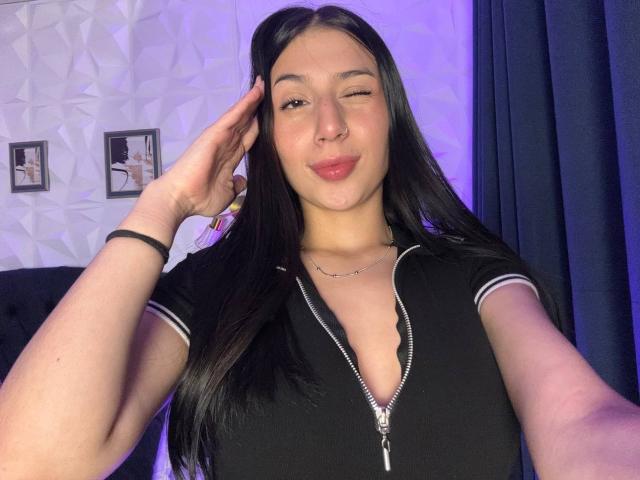 SophieBeaumont with long black hair, winking, wearing a black zip-up shirt, pink lips, hand near ear, blue curtain, purple light, and subtle jewelry. Boobs visible through zipper.