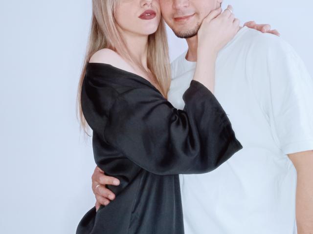 PoisonAndZeta, con lunghi capelli biondi, una camicia nera con una spalla scoperta e rossetto rosso, tiene sollevato il mento di un ragazzo con una camicia bianca, mentre lui le mette un braccio intorno, entrambi sorridono..