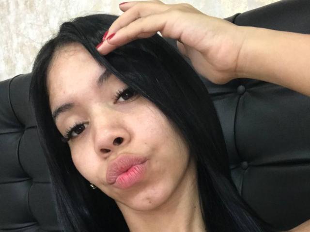 EmaGarces, avec ses longs cheveux noirs, ses lèvres roses et gonflées, et ses ongles rouges, tirant la langue, touchant ses cheveux avec sa main droite, assise sur un canapé en cuir noir..