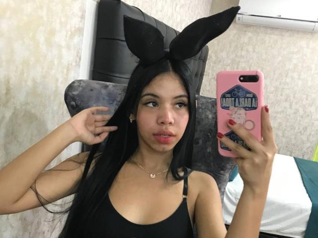EmaGarces avec des oreilles de lapin noires, de longs cheveux noirs, portant un débardeur noir, tenant un téléphone rose, montrant son rouge à lèvres et ses petites seins dans un selfie devant un miroir..