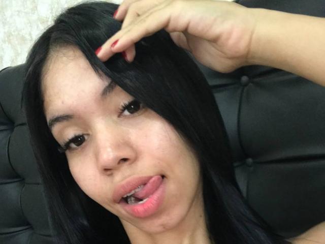 Ema Garces, com cabelo preto, a língua ligeiramente para fora, tocando o cabelo com unhas pintadas de vermelho, olhando para a câmera com olhos escuros, sentada em um sofá de couro preto..
