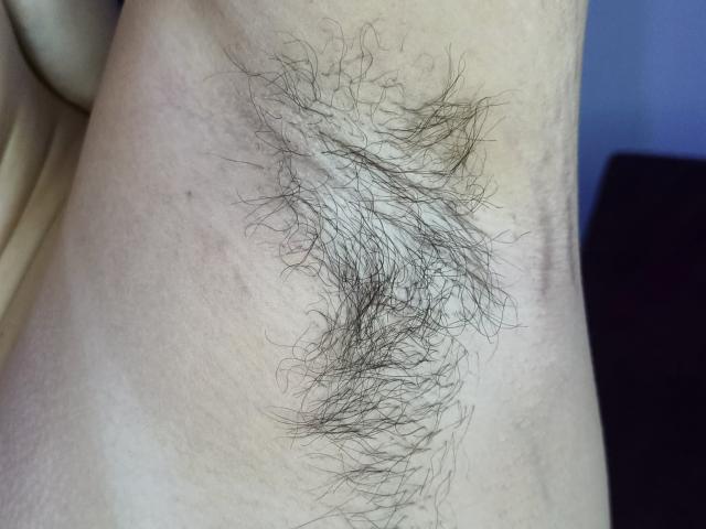 AlanisSweet's hairy armpit showing a thick, dark, curly patch of body hair against pale skin. Image focuses on the hairy area, with subtle skin texture visible.