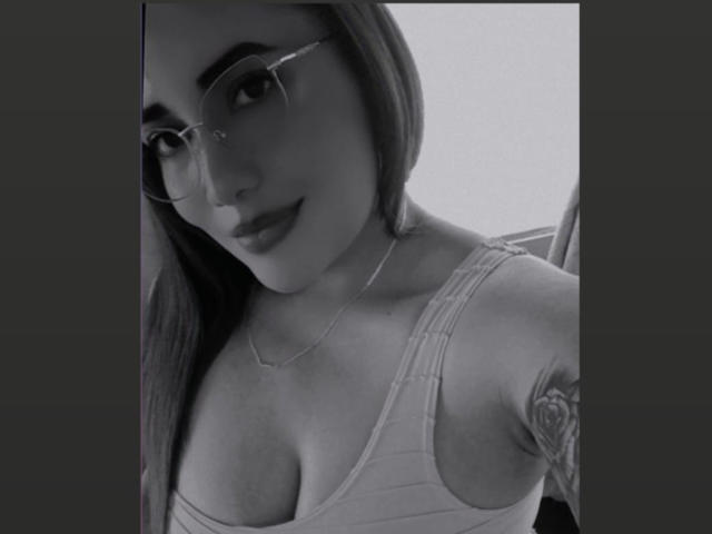 Black-and-white selfie of AlizWolf with glasses, a tattoo on her right arm, and a low-cut tank top showing her tits. She's smiling and has long hair.
