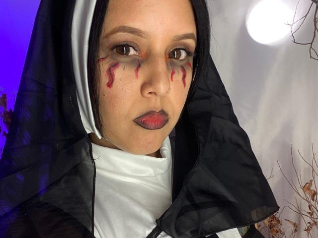Linger with black and white nun outfit, red makeup stains on face, black lipstick, and serious expression. Sheer black veil, white head covering, and subtle purple light.