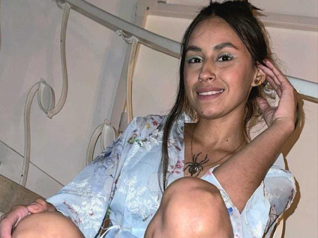 LizyGrey with brown hair, smiling, wearing a floral dress, has a spider tattoo on her chest, sits with knees up, hand in hair, white metal railing behind.