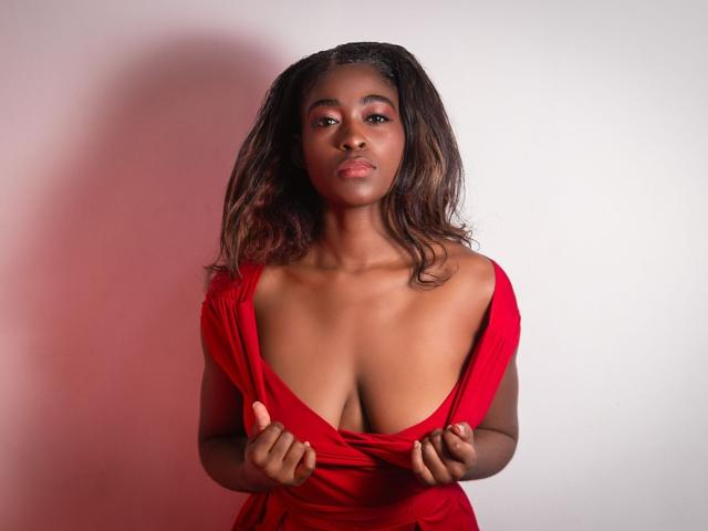 Scarlet Haylison, com pele escura, cabelo castanho ondulado e um vestido vermelho, está puxando a gola para baixo para expor seus seios grandes e arredondados. Ela tem sombra rosa nos olhos e uma expressão séria..