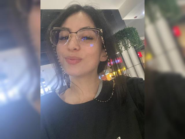 Camille Rios trägt eine Brille, hat dunkles Haar und ein leichtes Lächeln mit rosafarbenen Lippen. Sie trägt ein schwarzes Hemd und eine silberne Kette. Reflexionen von Lichtern sind auf ihrer Brille zu sehen..