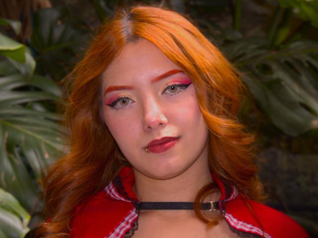 EmmaThomsonn has orange hair, pink eyeshadow, red lipstick, and a nose piercing. She's wearing a red shirt with a black choker. Her tits are not visible.