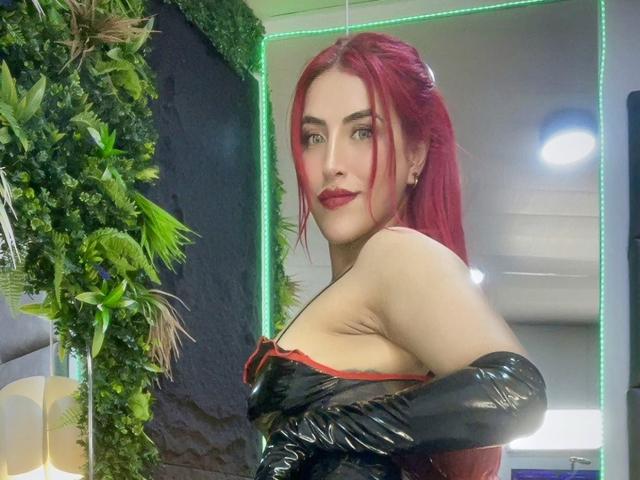 AlissaBrown with red hair, wearing black latex gloves and a red-trimmed black top, showing her tits, looking over her shoulder with a confident expression.