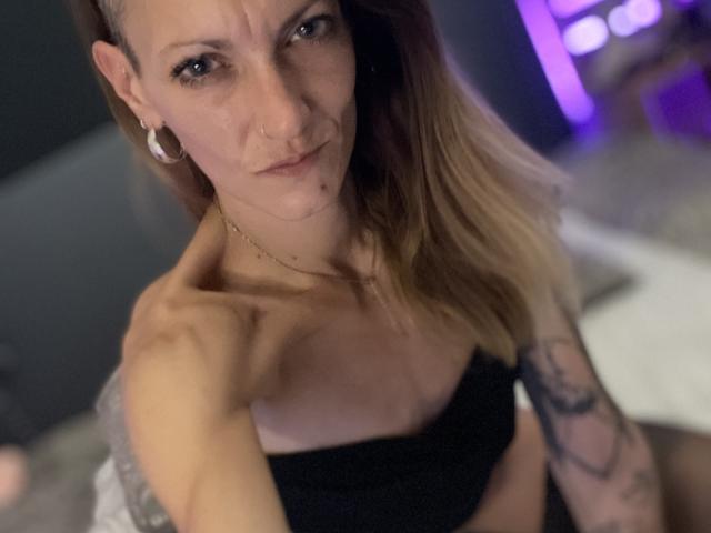 DaneeSoHot con un lado de la cabeza rapado, pechos pequeños, un top negro sin tirantes, un brazo tatuado, aros grandes, un piercing en la nariz y cabello rubio, mirando hacia arriba con una expresión ligeramente molesta en una habitación con poca luz..