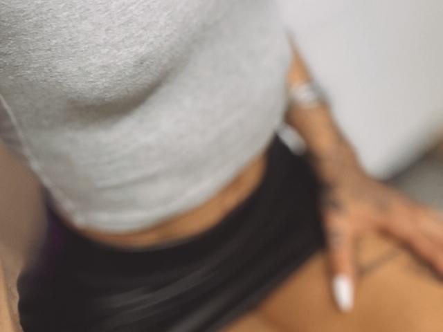 DaneeSoHot's small, tight, gray shirt showing her tits, black skirt, tan skin, tattooed hand with white nails, and a watch on her wrist in a blurred, shallow-depth image.