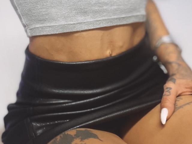 DaneeSoHot's toned midriff with a visible navel, wearing a black leather skirt, gray crop top, and tattooed hand with white nails. Her tan skin contrasts with the black skirt.