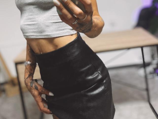 DaneeSoHot indossa una canotta grigia che mette in risalto i suoi addominali scolpiti, una gonna in pelle nera, ha dei tatuaggi sulle mani, tiene un telefono e l'attenzione è concentrata sulla parte inferiore del corpo..