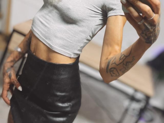 O corpo tonificado e bronzeado de DaneeSoHot, vestindo uma blusa cinza justa e uma saia de couro preta. Tatuagens visíveis nos braços, segurando um telefone e usando anéis..