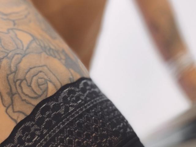 O lado tatuado de DaneeSoHot, com uma grande rosa, calcinhas de renda preta e o braço tatuado, em uma luz suave. Suas nádegas estão parcialmente visíveis, destacando o padrão da renda..
