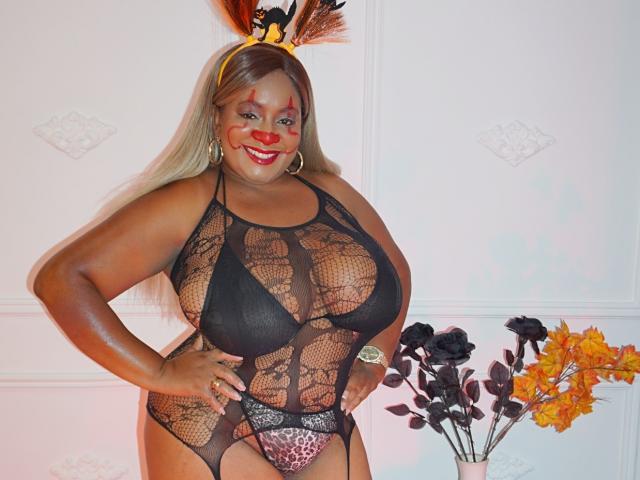 Nahomi Anders, com seios grandes, pele escura, vestindo um macacão de renda preta, calcinha de estampa de leopardo, e uma tiara de penas, sorrindo, com as mãos nos quadris, maquiagem vermelha no rosto e flores laranjas..