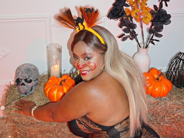 Nahomi Anders mit orangefarbenen Federn im Haarreif, lächelnd, mit geröteter Wange, trägt eine schwarze Spitzenbluse, orangefarbene Kürbisse, einen Schädel, eine Kerze und schwarze Rosen in einer Vase auf Heu..