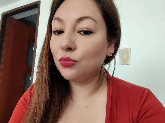 SakotoDika hat helle Haut, braunes Haar, roten Lippenstift und zieht die Lippen zusammen, wobei ihre Augen halb geschlossen sind. Sie trägt ein rotes Oberteil und große Ohrringe..
