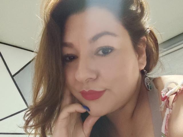 SakotoDika a les lèvres rouges, un vernis à ongles bleu, les cheveux bruns et un joli visage avec un léger sourire. Elle porte des bretelles florales et grises. Sa main est sur sa joue..