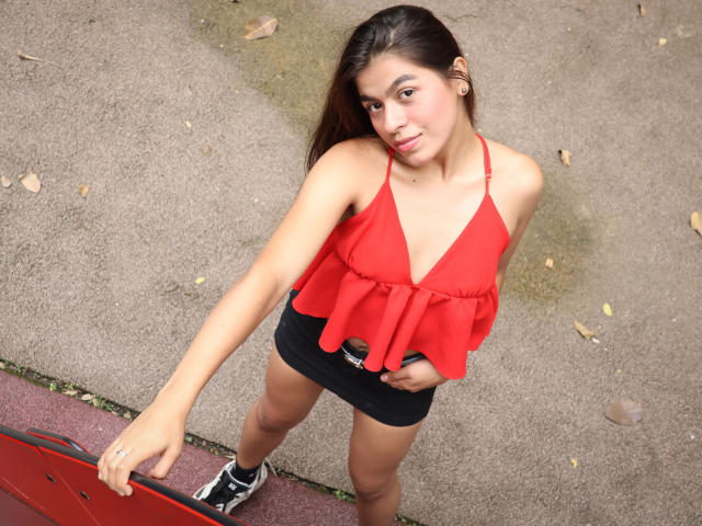 MaiiaWest indossa una maglietta rossa, pantaloncini neri e scarpe da ginnastica nere, e tiene in mano una struttura da gioco rossa. Ha un seno di media grandezza, capelli castani e un leggero sorriso, e guarda verso la fotocamera..