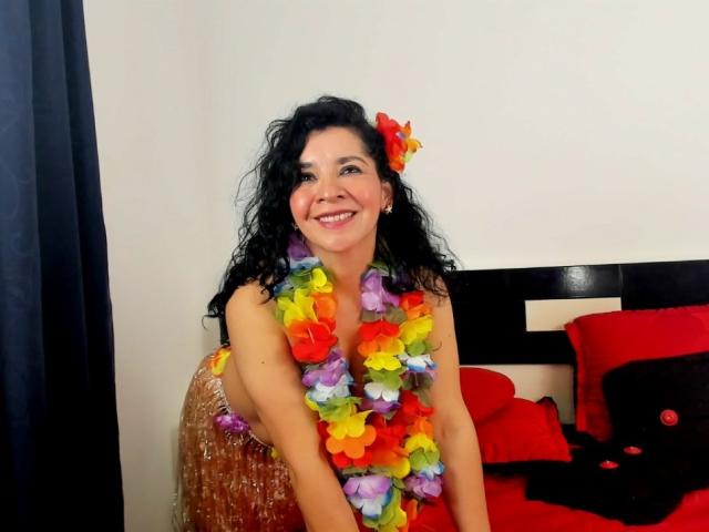 EtoileLatin, a curvy woman with dark curly hair, wearing a colorful flower lei, smiling, in a red dress, standing by a black and white bed with red pillows.