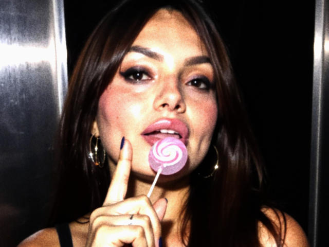 GalaMalkova with dark brown hair, holding a pink and white swirled lollipop near her slightly open, glossy pink lips, wearing gold hoop earrings, and dark nail polish.