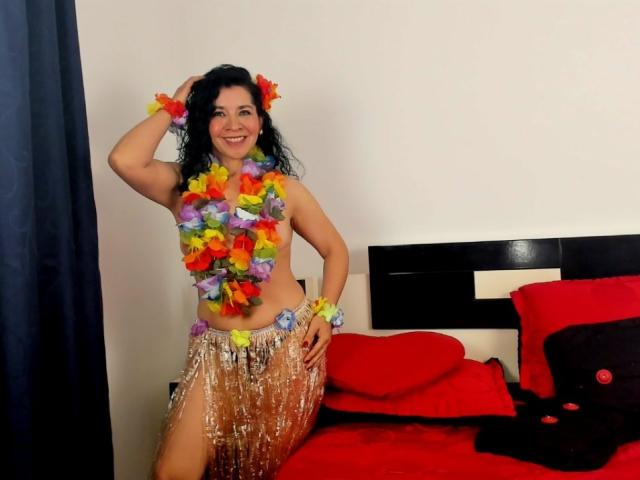 EtoileLatin with curly black hair, smiling, wearing a colorful flower lei, and a silver sequin skirt, exposing her tits. She's standing next to a red bed with black pillows.