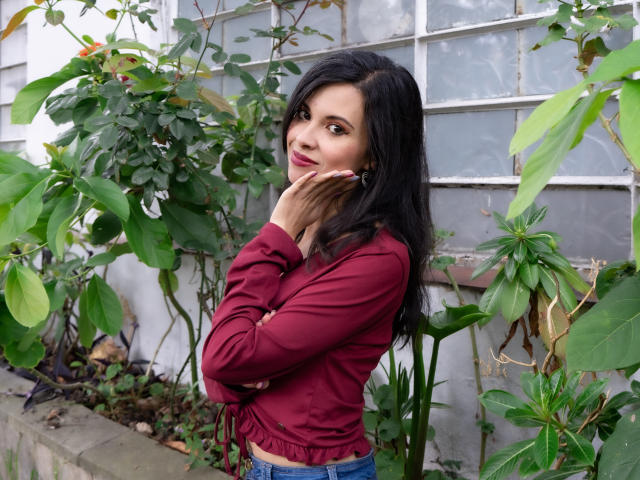TaylaCooperk with long black hair, wearing a red long-sleeve top, hands near her face, looking slightly to the side, standing among green plants. Her tits are not visible.