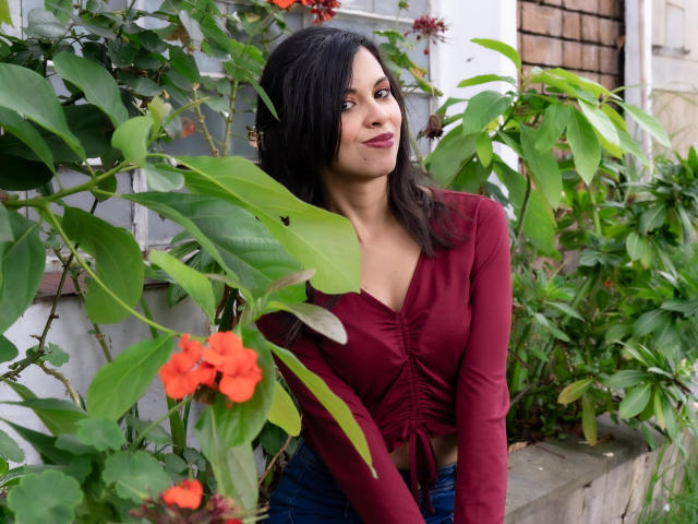TaylaCooperk with dark hair, red top, and blue jeans, standing in front of green plants with orange flowers, smiling slightly, looking to the side.