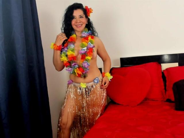 EtoileLatin with curly black hair, wearing a colorful flower lei, red heart pillows, and a silver sequined skirt, smiling, showing her bare tits, and standing by a red bed.