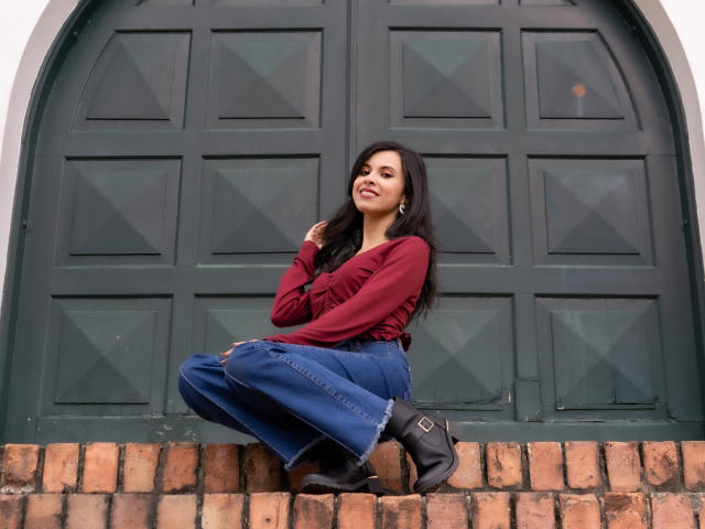 TaylaCooperk has long black hair, wears a red long-sleeve top, blue flared jeans, black boots, and sits on brick steps in front of a green arched door.