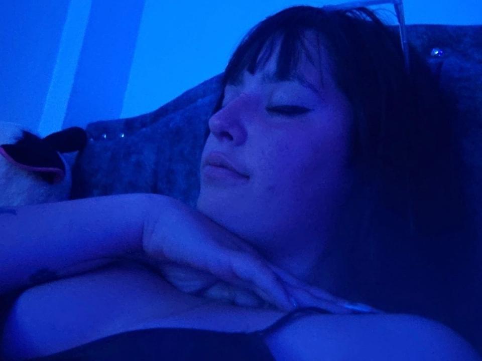 Katsumi69 with blue-tinted skin, eyes closed, resting head on hand, showing small boobs, slight smile, dark brown hair, and a tattoo on her right hand.