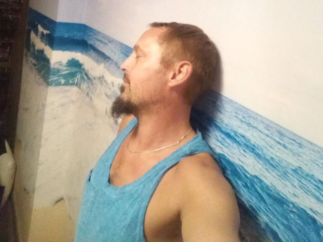 A photo of Major-hot with a beard, short brown hair, and a blue tank top, leaning against a wall with a blue wave pattern. His ass is not visible.