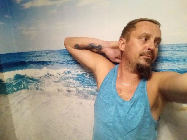 Major-hot with a blue tank top, tattoo on right arm, short brown hair, beard, and necklace, posing with arms behind head, looking to the side, in a beach-themed setting.