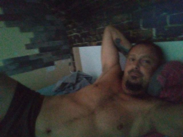 Major-hot, shirtless, with a beard and tattoo, lies on a bed, arms behind head, wearing dark underwear, looking at the camera with a relaxed, slightly goofy expression.